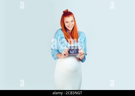 Bonne adorable fille dans l'espérance tenant la photo de l'écran de bébé et souriant merveilleusement à l'appareil photo isolé sur fond bleu clair. Modèle de race mixte, Banque D'Images