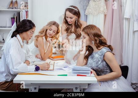 groupe caucasien de femmes se sont rassemblées dans le bureau de designer pour discuter de nouveau style de robe, les jeunes filles dans les robes de mariage blanches tendance Banque D'Images