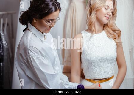 belle femme caucasienne client et tailleur dans la boutique de mariage, prenant des mesures du corps pour la création nouvelle robe, la préparation d'un mariage Banque D'Images