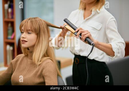 coiffeur professionnel faisant coiffer la jeune fille caucasienne avec les cheveux longs, coiffeur prudent et jeune beau client dans le salon de beauté Banque D'Images