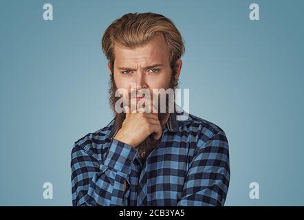 Frognant homme pensant exprimant des doutes et des préoccupations. Petit garçon avec barbe en chemise à carreaux bleu isolée sur fond bleu studio. Negat Banque D'Images