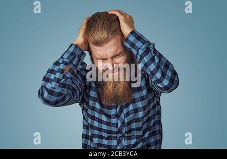 Homme déprimé ayant un mal de tête très fort. Petit garçon avec barbe en chemise à carreaux bleu isolée sur fond bleu studio. Face négative Banque D'Images