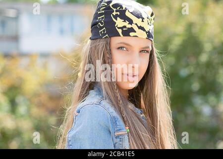 Jolie femme moderne adolescente avec de longs cheveux portant bandana et regarder sérieusement l'appareil photo en plein soleil Banque D'Images