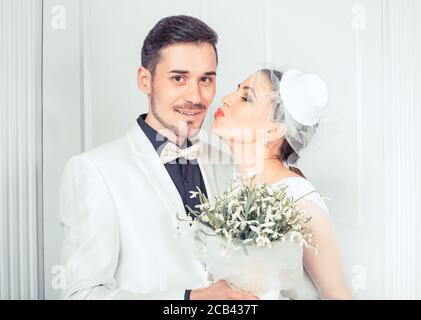 Belle jeune femme en robe de mariée et voile embrassant beau gustain dans la joue debout dans l'étreinte Banque D'Images