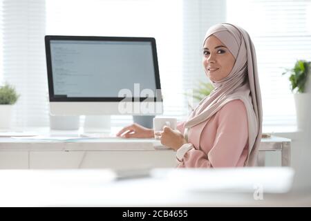 Portrait d'une jeune musulmane souriante dans un hijab assis bureau avec ordinateur et café dans le bureau Banque D'Images