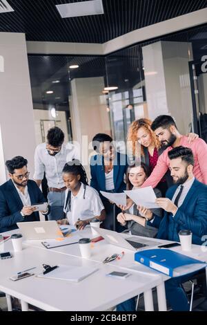 Groupe d'excités gaie divers collaborateurs dans l'usure formelle tableau surround avec ordinateur portable, un café à emporter et des feuilles de papier et des dossiers de travail, Banque D'Images