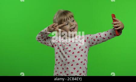 Enfant fille utilisant un smartphone. Portrait d'un enfant aux cheveux blonds émotionnellement fait le selfie sur le téléphone mobile. Appel vidéo, blog, jeux, regarder un téléphone portable. Montrer des visages amusants. Fond Chroma Key Banque D'Images