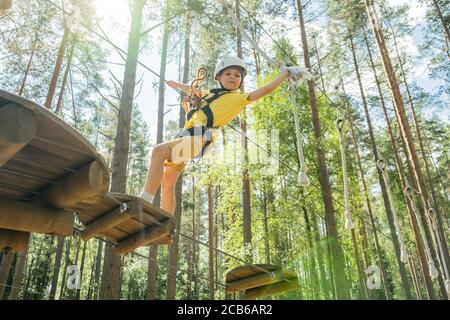 Petit garçon avec équipement d'escalade piste de corde d'escalade entre les pins dans un parc d'aventure. Parc à cordes. Bonne journée d'été. Banque D'Images