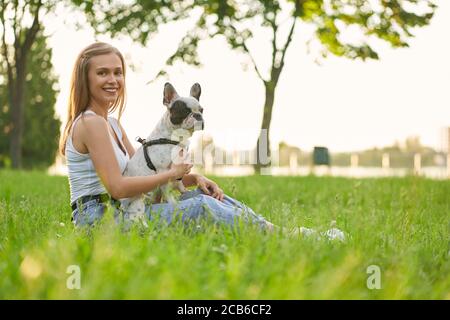 Vue latérale de la jeune femme heureuse assise sur l'herbe, tenant le joli boudogue français sur les jambes. Magnifique fille souriante profitant du coucher du soleil d'été, chien d'animal dans le parc de la ville et regardant l'appareil photo. Banque D'Images