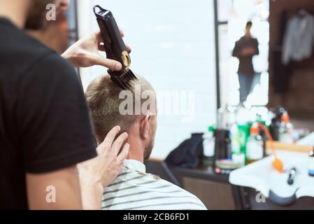 Vue rapprochée arrière d'un homme au salon de coiffure ayant une coupe de cheveux tendance. Un coiffeur méconnaissable portant un t-shirt noir travaille avec une tondeuse noire dans sa main Banque D'Images