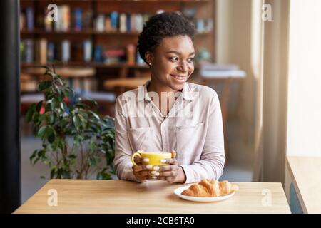 Bonne fille noire avec une tasse de café frais et délicieux croissant à la table dans le café Banque D'Images