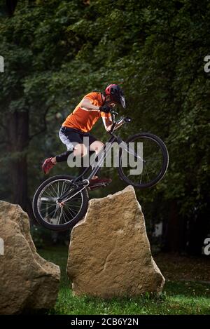 Vue de face d'un homme tenant un guidon de vélo et faisant une cascade sur des pierres. Athlète essayant d'atterrir sur le vélo de montagne sur l'herbe. Concept de terrain. Banque D'Images