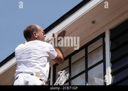 Peintre de maison professionnel debout sur échelle et ponçage du bois de la gouttière d'un dormeur en préparation pour la peinture Banque D'Images