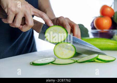 Gros plan sur les mains des femmes qui tiennent le couteau et coupent la courgette fraîche sur le plan de coupe. La main femelle coupe le concombre japonais cru en cercles avec un couteau sur cu Banque D'Images