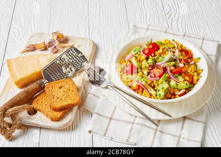 salade de maïs sucré avec avocat, tranches d'oignon rouge et tomates, nappées de fromage râpé dans un bol blanc sur une table en bois, vue sur le paysage depuis le dessus Banque D'Images