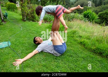 Père allongé sur l'herbe du sol dans le jardin de campagne jouant avec l'équilibre de son enfant de 3 ans le tenant dedans Air sur les jambes surélevées pays de Galles Royaume-Uni KATHY DEWITT Banque D'Images