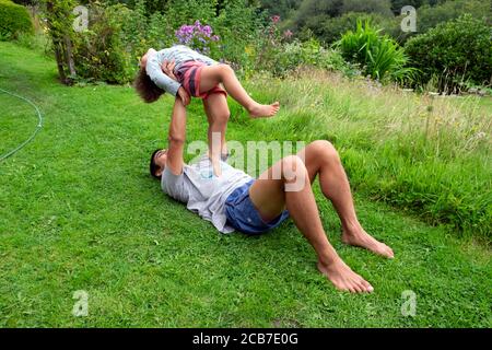 Père allongé sur l'herbe du sol dans le jardin de campagne jouant avec l'équilibre de son fils de 3 ans enfant garçon tenue Lui dans l'air pays de Galles Royaume-Uni KATHY DEWITT Banque D'Images