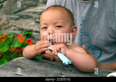 Bébé de race mixte tenant une cuillère en plastique assis à l'extérieur manger dîner à une table de pique-nique organisée par le père Été pays de Galles Royaume-Uni KATHY DEWITT Banque D'Images