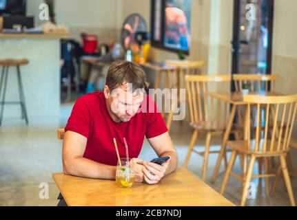 Jeune sérieux pensif attentionné homme tendu penser envoyer un message par smartphone dans une chemise rouge dans un café boire du thé de glace chia. De vraies personnes Banque D'Images