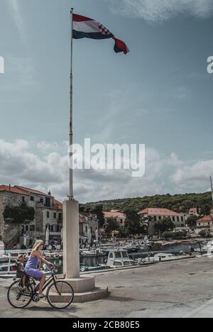 Postira, Croatie août 2020 Maman vélo avec un enfant autour d'un grand poteau avec le drapeau croate sur lui. Journée ensoleillée près du port de la vieille ville Banque D'Images