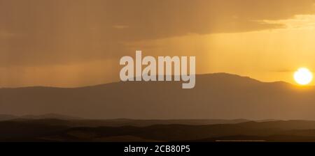 Paysage montagneux au coucher du soleil avec un rideau de pluie éloigné à l'horizon et le disque solaire se cachant Banque D'Images