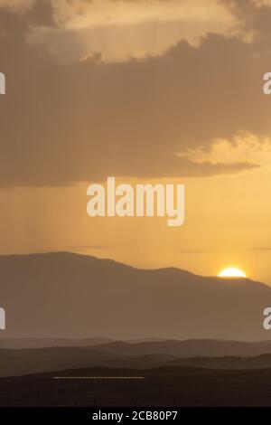 Paysage montagneux au coucher du soleil avec un rideau de pluie éloigné à l'horizon et le disque solaire se cachant Banque D'Images