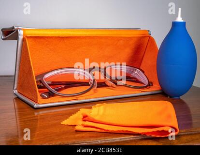 Gros plan d'une paire de lunettes dans un boîtier de rangement de protection rigide orange au design moderne, avec souffleur de poussière en caoutchouc bleu et chiffon de nettoyage orange à proximité. Banque D'Images