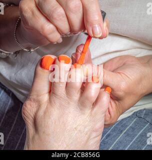 Une main tenant le pied d’une vieille femme, tout en utilisant un pinceau pour peindre un ongle, pour compléter les cinq en peinture orange vif pour les ongles. Banque D'Images