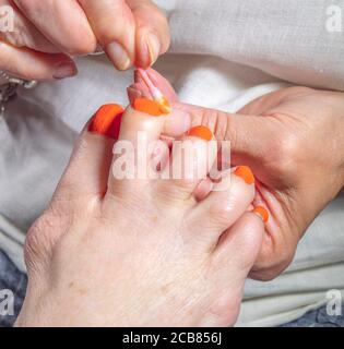 Une main tenant le pied d’une vieille femme, tout en utilisant un coton-tige pour nettoyer l’excès de peinture autour d’un ongle, après avoir peint les cinq en orange vif. Banque D'Images