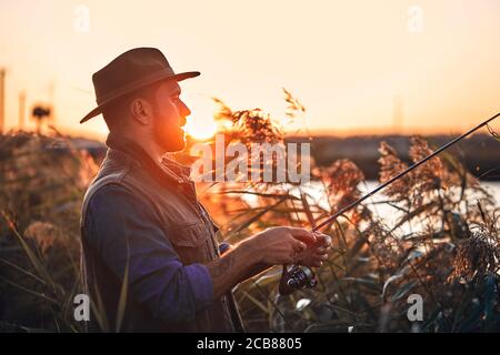 Homme barbu caucasien dans la pêche au chapeau en roseau près du lac. Coucher de soleil. Banque D'Images