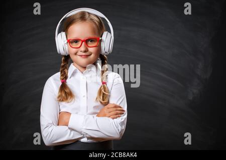 Gros plan d'une petite fille adorable élève dans un casque et des lunettes sur le fond d'un tableau noir d'école. Un enfant sérieux, intelligent et confiant, a traversé la frontière Banque D'Images