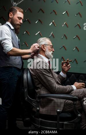 Coiffure brosses les cheveux gris de l'homme respectable client habillé en tailleur dans le salon de coiffure. Beau salon de coiffure, nouvelle coiffure alors qu'un vieux riche Banque D'Images