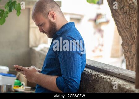 Homme à barbe chauve portant une chemise bleue avec des points inclinés sur un mur en pierre, tenant un téléphone et le regardant Banque D'Images
