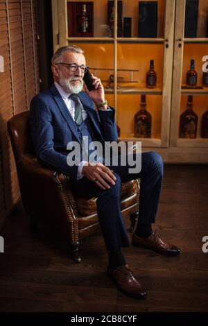 Vieux médecin privé expérimenté en lunettes et costume élégant chic chic en conversation sur smartphone avec son patient tout en se reposant dans le grand bras mou- Banque D'Images