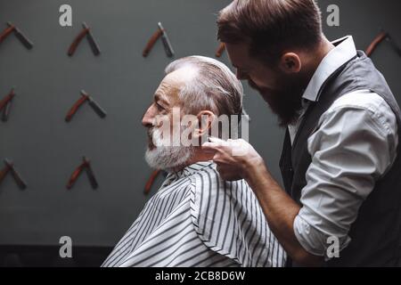 Les jeunes professionnels de coiffure en donnant une coupe avec la concentration d'un riche gentleman respectable, debout et tenant en rasoir client âgé détendue Banque D'Images