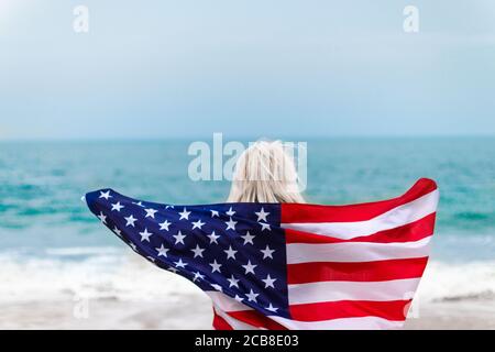 Femme blonde caucasienne tenant le drapeau américain en arrière et regardant mer Banque D'Images