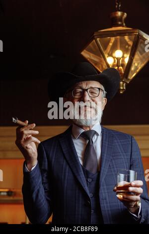 Élégant homme vieux avec chapeau à large bord et costume bleu foncé pour hommes fumant cigare intérieur, debout près du comptoir du bar avec boissons alcoolisées. Et toilettes Banque D'Images