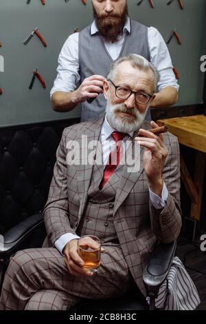 Les jeunes professionnels de coiffure en donnant une coupe avec la concentration d'un riche gentleman respectable, debout et tenant en rasoir client âgé détendue Banque D'Images