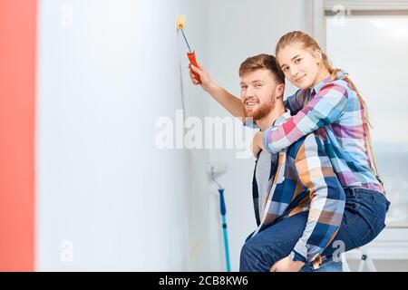 Homme à barbe gai donnant une promenade en pigeyback à sa femme alors qu'elle peint le mur avec une brosse, tout en rénovant l'appartement Banque D'Images