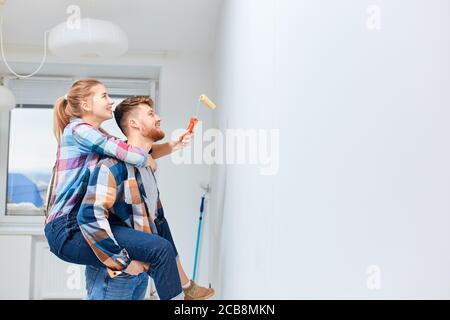 Homme à barbe gai donnant une promenade en pigeyback à sa femme alors qu'elle peint le mur avec une brosse, tout en rénovant l'appartement Banque D'Images