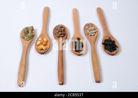 Différentes graines - maïs, courge, café, poivre, riz, tournesol - en cuillères en bois sur une surface blanche Banque D'Images