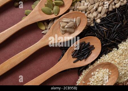Différentes graines - quinoa, potiron, café, poivre, riz, tournesol - en cuillères en bois sur une surface de différentes graines et fond brun Banque D'Images