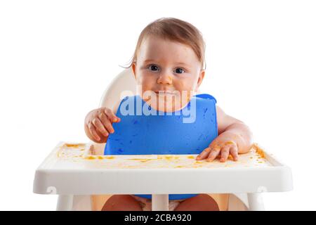 Bébé gai enfant mange lui-même de la nourriture avec les mains. Portrait d'un garçon d'enfant sale heureux en chaise haute et désordonné autour. Banque D'Images