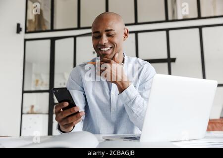 Photo de rire de l'homme afro-américain en utilisant le téléphone mobile pendant travailler avec un ordinateur portable à la table dans le salon Banque D'Images