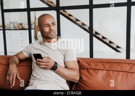 Photo d'un homme afro-américain qui pense en utilisant un téléphone portable tout en étant assis sur le canapé dans la salle de séjour Banque D'Images
