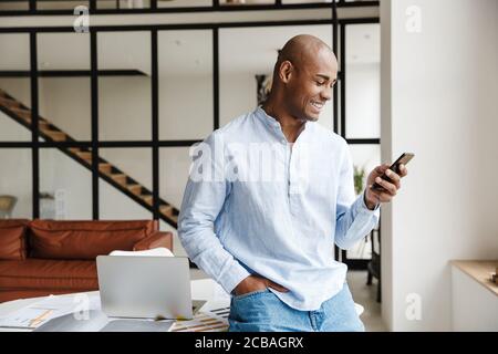 Photo de rire de l'homme afro-américain en utilisant le téléphone mobile pendant travailler dans le salon à la maison Banque D'Images