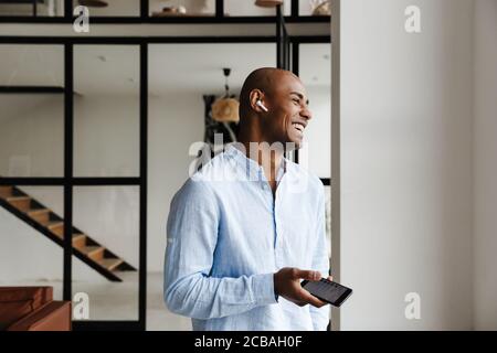 Photo d'un homme afro-américain joyeux qui rit tout en utilisant le sans fil écouteurs et téléphone portable dans le salon Banque D'Images