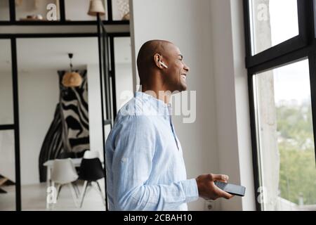 Photo d'un homme afro-américain joyeux qui rit tout en utilisant le sans fil écouteurs et téléphone portable dans le salon Banque D'Images