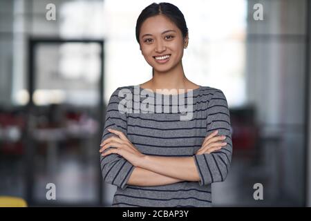 Portrait d'une jeune femme asiatique heureuse, employée de bureau dans des vêtements décontractés en gardant les bras croisés, en regardant l'appareil photo et en souriant tout en restant debout Banque D'Images