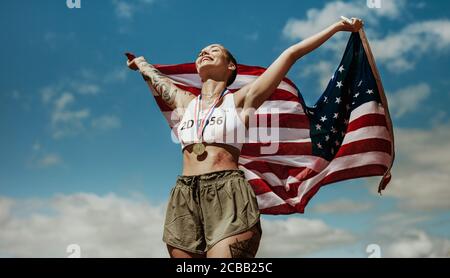 Athlète bénéficiant de la victoire avec le drapeau national américain contre le ciel. Femme coureuse avec des médailles célébrant une victoire tenant le drapeau américain. Banque D'Images